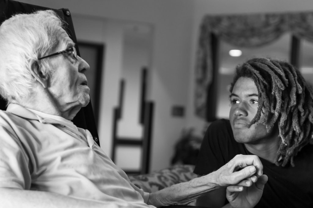 a young Black man with dreadlocks and black t-shirt is softly looking at and holding the hand of his grandfather, an elder white man with short white hair and glasses wearing a light gray shirt.