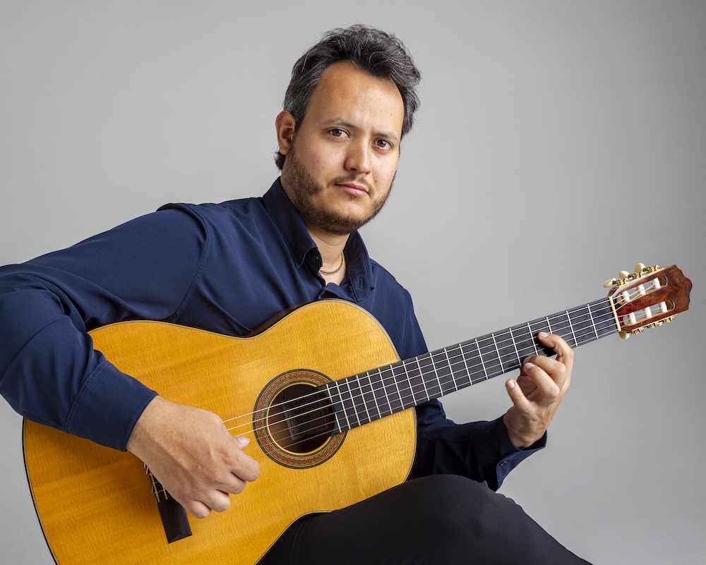 Misael, a man with light brown skin wearing a blue collared long-sleeve shirt, holding a guitar and staring at the viewer