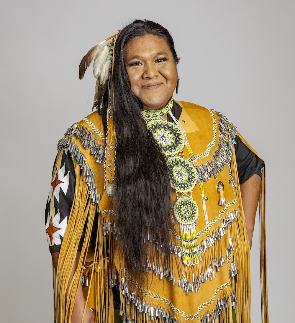 Mylyle, a smiling White Mountain Apache woman wearing a buckskin dress with feathers in her long, dark hair.