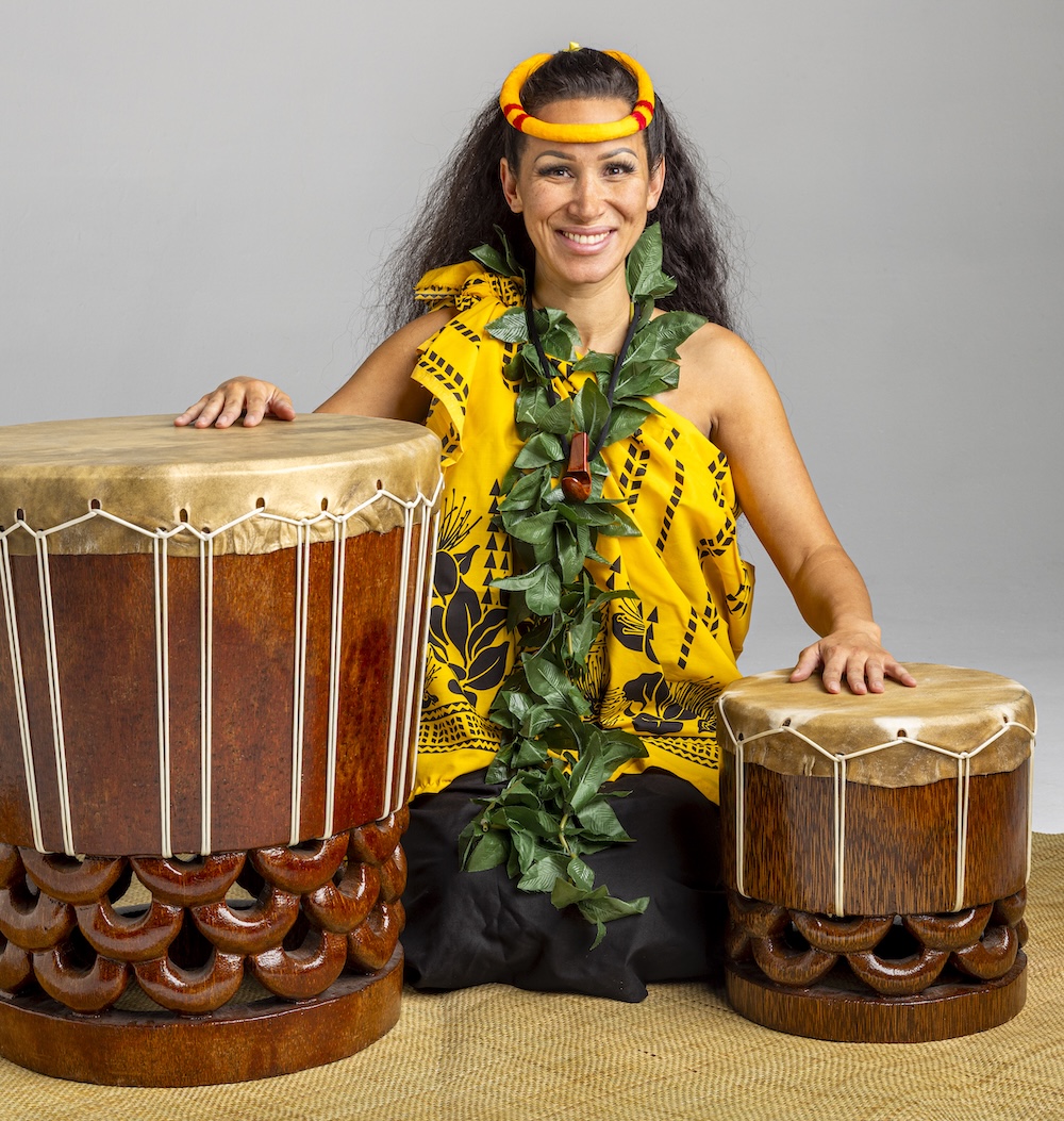 Nālani, a descendant of Native Hawaiians, is dressed in a yellow dress and headband posing with her hands on drums.