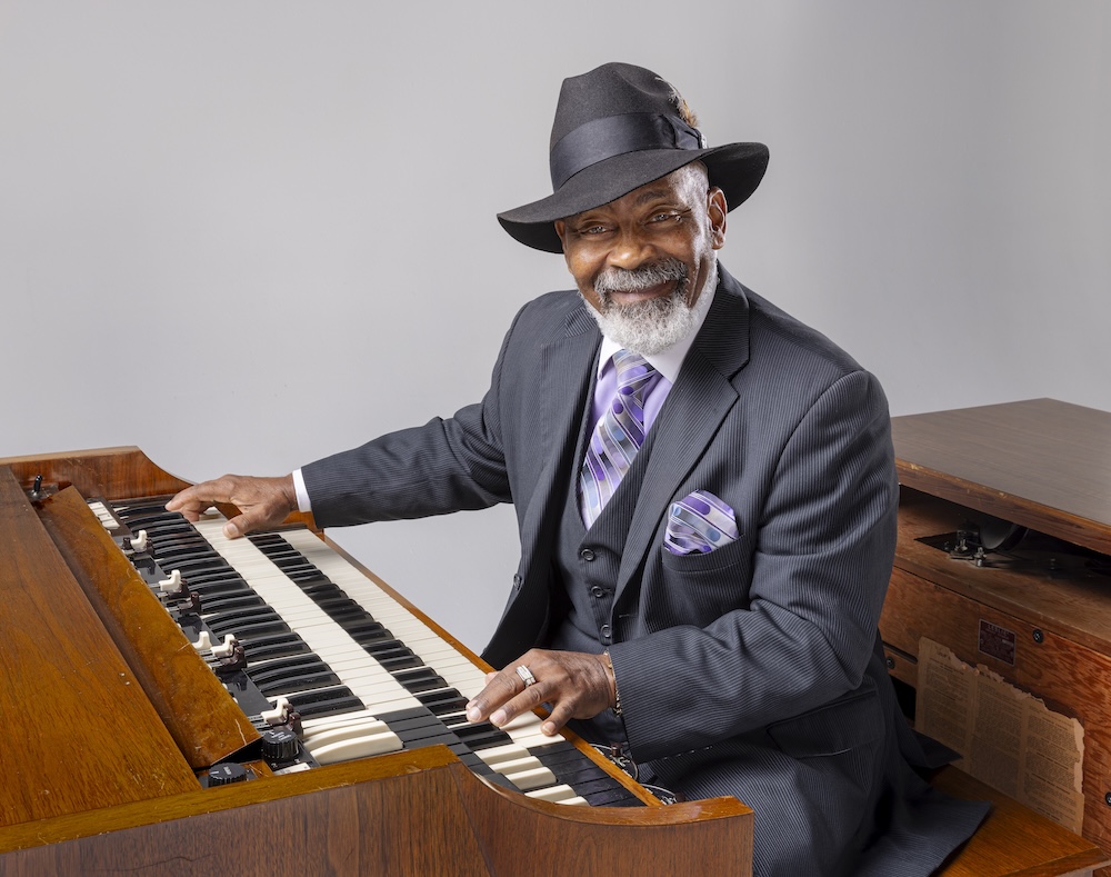 Royce, a Black elder wearing a dark gray suit with matching hat and a purple tie and pocket square, is playing the organ.